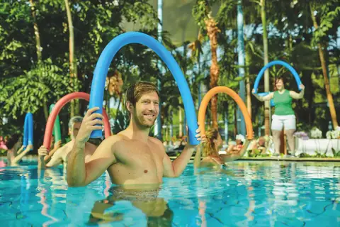 Mann mit blauer Poolnudel bei Wassergymnastik im tropischen Hallenbad mit Palmen