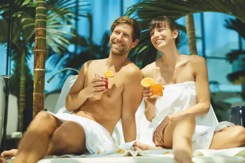 Pärchen in Bademänteln mit Cocktails auf Liegen in tropischer Saunalandschaft
