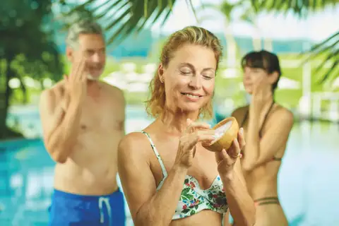 Frau mit geschlossenen Augen hält Holzschale mit Creme, zwei Personen im Hintergrund am Pool