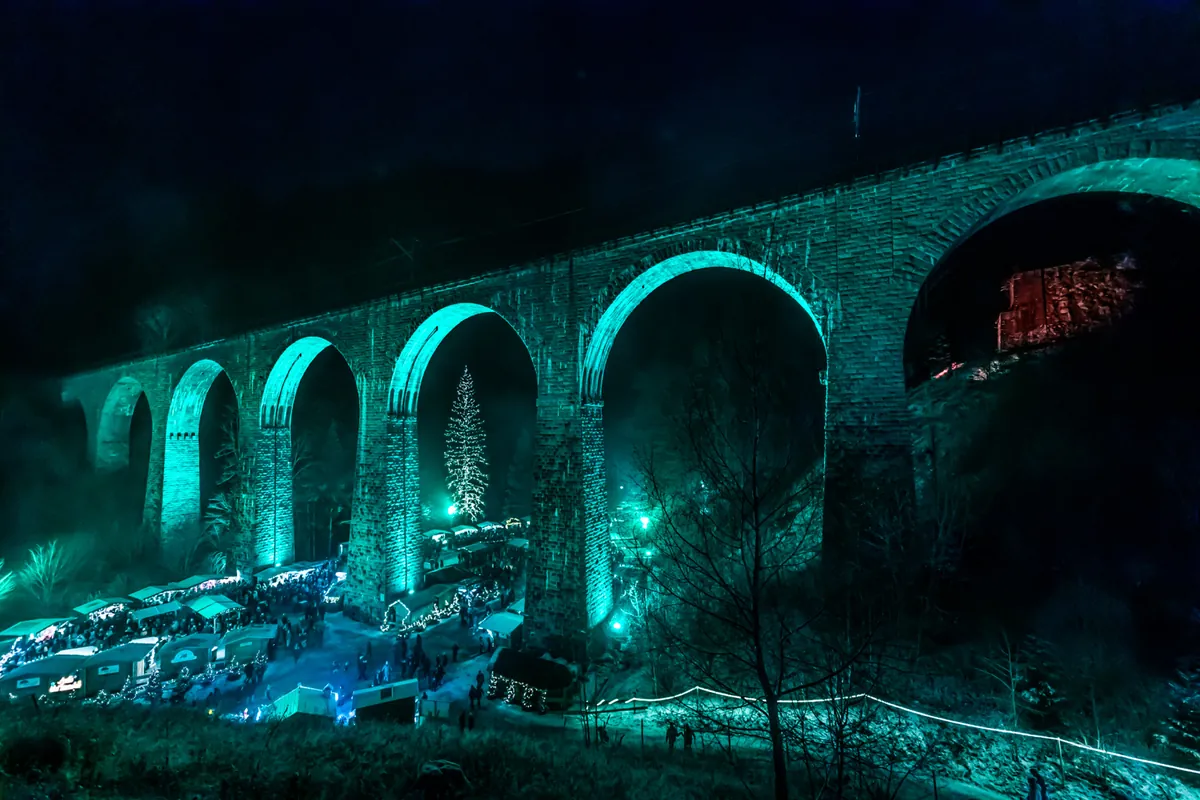 Ravenna-Viadukt bei Nacht in türkis beleuchtet, darunter beleuchteter Weihnachtsmarkt mit Besuchern