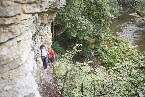 Zwei Wanderer auf schmalem Pfad an Felswand entlang der Wutach, umgeben von üppigem Grün