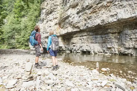 Zwei Wanderer mit Rucksäcken stehen am Flussufer der Wutachschlucht vor mächtiger Kalksteinwand