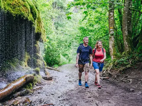 Zwei Wanderer auf einem Waldweg neben moosbewachsenem Felsen mit Wasserfall, Schwarzwald