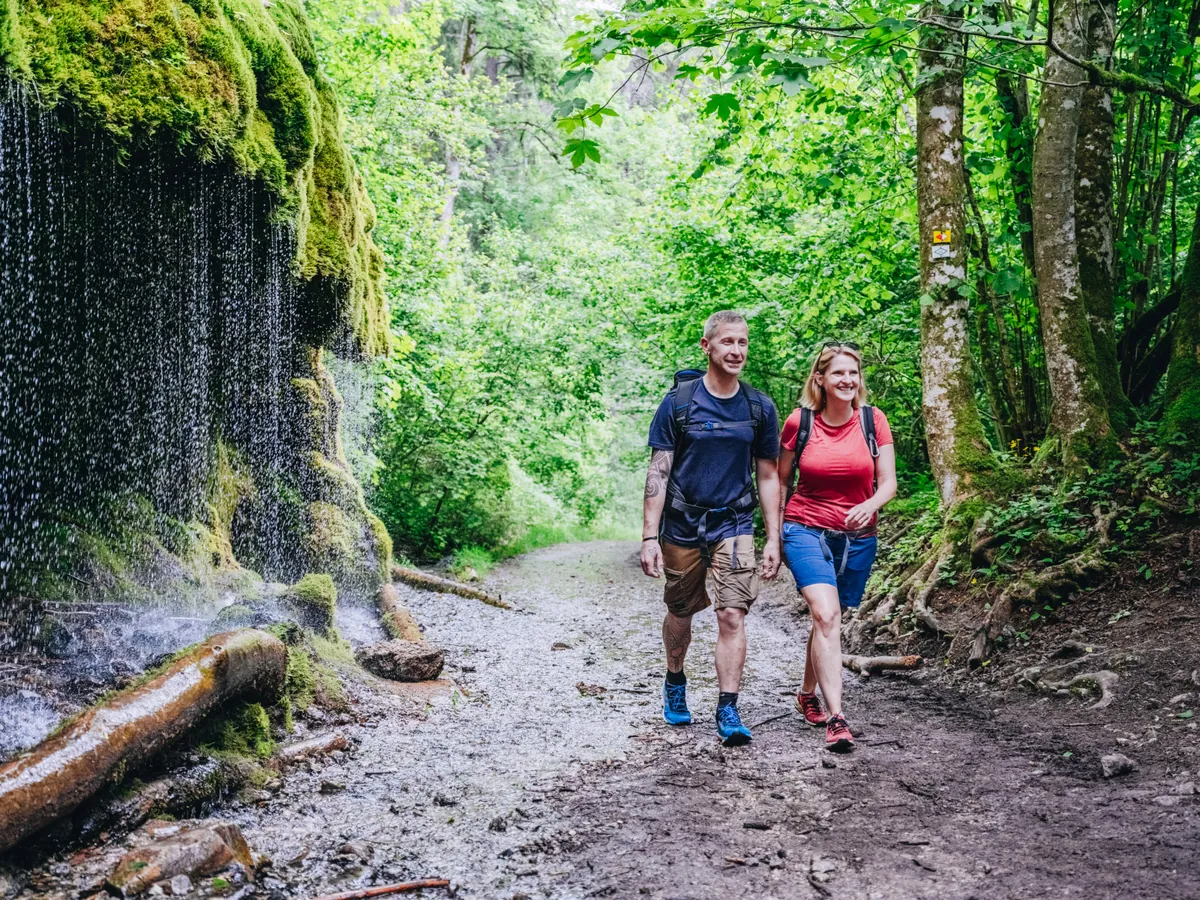 Zwei Wanderer auf einem Waldweg neben moosbewachsenem Felsen mit Wasserfall, Schwarzwald