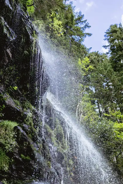Wasserfall stürzt über bemooste Felswand, umgeben von grünen Bäumen und blauem Himmel