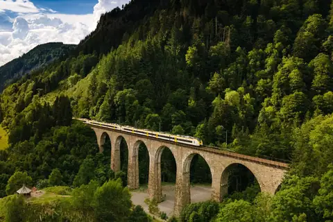 Zug fährt über steinernen Eisenbahnviadukt durch bewaldete Berglandschaft