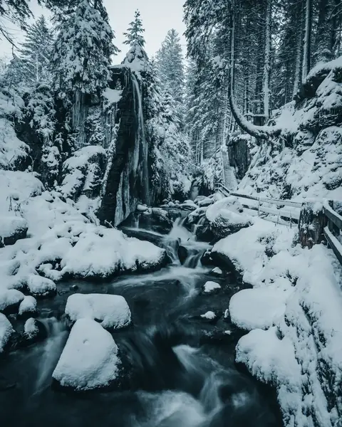 Menzenschwander Wasserfall im Winter, vereist und verschneit, mit Holzsteg und Bachbett im Vordergrund