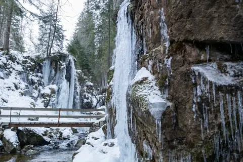 Gefrorener Wasserfall mit Eisvorhang und verschneiter Holzbrücke im Schwarzwald, winterliche Schlucht