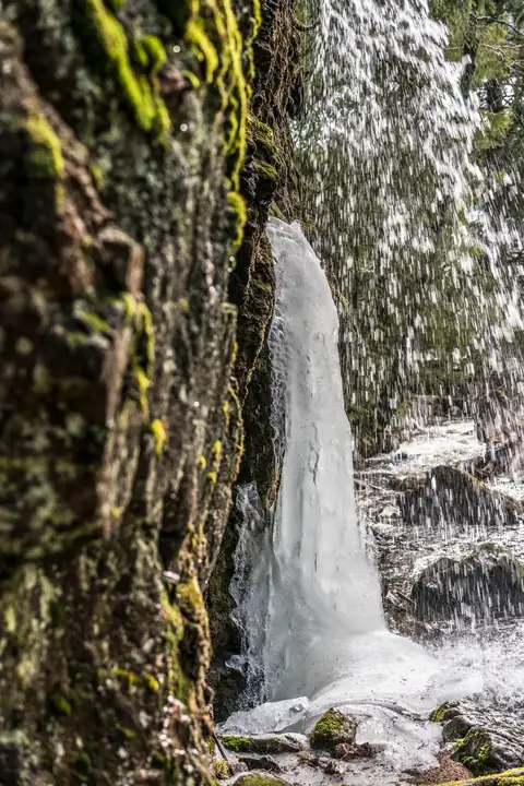 Menzenschwander Wasserfall im Winter: Eiszapfen vor moosiger Felswand, dahinter stürzendes Wasser