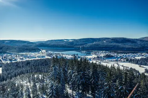 Winterlicher Blick von einem Aussichtspunkt über Nadelwald auf einen See und ein verschneites Dorf im Schwarzwald