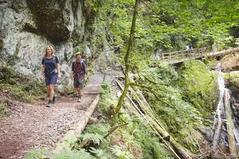 Zwei Wanderer auf schmalem Holzsteg durch bemoostes Schluchtgelände mit Wasserfall und Felswand