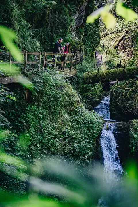 Paar auf Holzbrücke über Wasserfall in üppig begrünter Schlucht der Lotenbachklamm