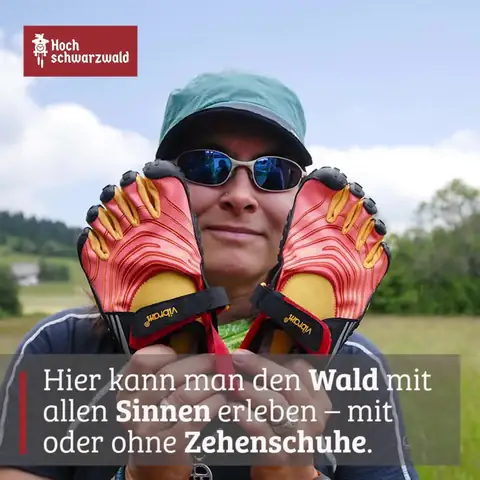 Person mit Basecap und Sonnenbrille haelt zwei rote Zehenschuhe vor das Gesicht, im Hintergrund Wiesenlandschaft
