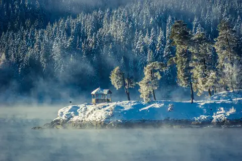 Nebelverhangenes Seeufer mit schneebedecktem Pavillon und Bäumen im Raureif