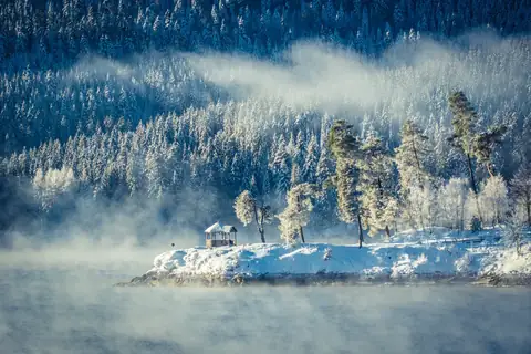 Nebliges Seeufer im Winter mit verschneiter Landspitze, Pavillon und schneebedeckten Nadelbäumen vor bewaldetem Hang