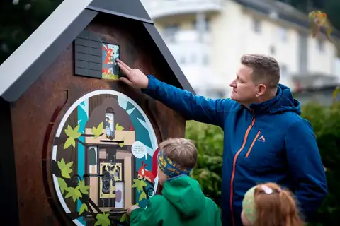Rostfarbenes Kuckucksuhr-Holzhaus mit Zahnrad-Illustration am Kuckucksuhrenpfad, Vater und Kinder schauen zu