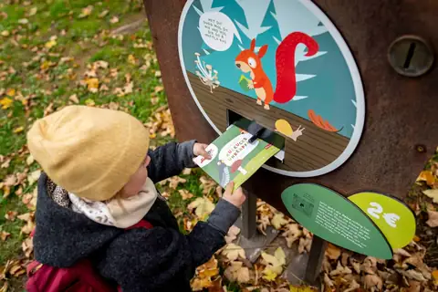 Kind wirft Wanderheft in eine bemalte Stationsbox des Kuckucksuhrenpfads mit Eichhoernchen-Illustration