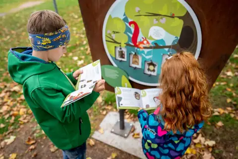 Kinder mit Aufgabenheft vor Infotafel mit Eichhörnchen und hängenden Kuckucksuhren am Waldweg