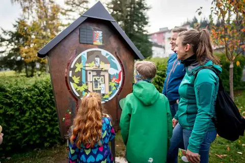 Familie mit Kindern betrachtet eine kunstvoll gestaltete Kuckucksuhr-Station am Wegesrand im Wald