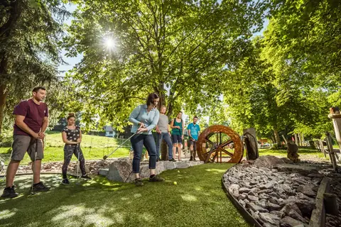 Gruppe spielt Adventure-Minigolf im Wald, Frau schlaegt Ball auf Kunstrasen-Bahn mit Wasserrad
