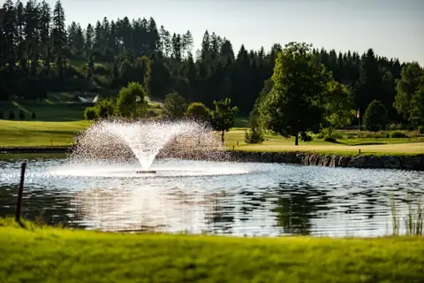 Golfplatz mit Wasserfontäne in einem Teich, umgeben von Grün und Wald