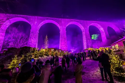 Lila beleuchtetes Eisenbahnviadukt über dem winterlichen Weihnachtsmarkt Ravennaschlucht bei Nacht