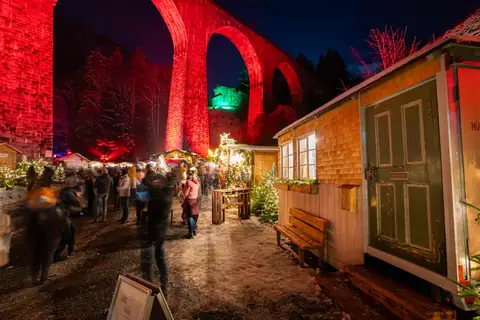 Nachts rot beleuchtetes Ravenna-Viadukt über dem Weihnachtsmarkt in der Ravennaschlucht mit Besuchern.