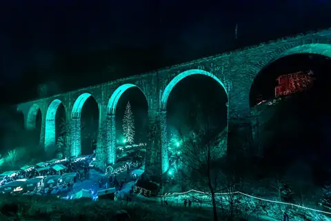 Türkisfarben beleuchtetes Eisenbahnviadukt bei Nacht über Weihnachtsmarktstaenden mit Christbaum