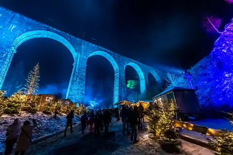Blau beleuchteter Eisenbahnviadukt bei Nacht über einem Weihnachtsmarkt mit Buden, Lichterbäumen und Besuchern im Schnee