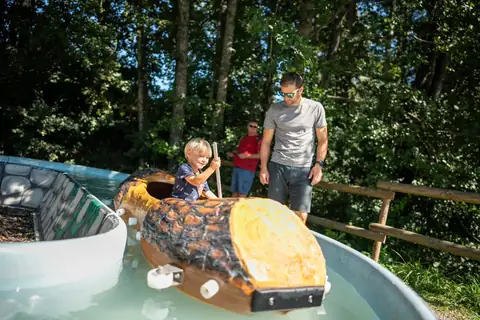 Kleinkind in Schildkröten-Fahrzeug auf Wasserfahrt, begleitet von Vater, grüne Kulisse
