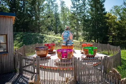 Tassen-Karussell mit bunten Tassen und Elefanten-Figur in der Mitte, umgeben von Holzzaun im Freien