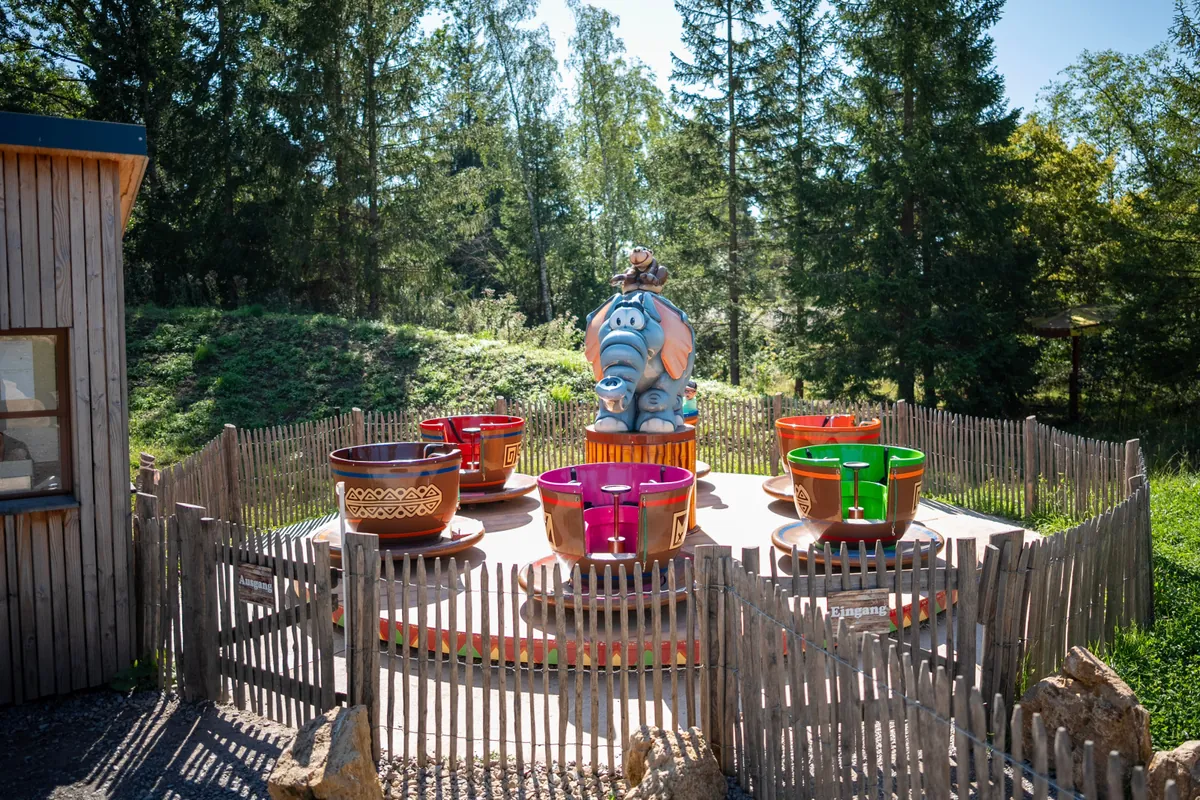 Tassen-Karussell mit bunten Tassen und Elefanten-Figur in der Mitte, umgeben von Holzzaun im Freien