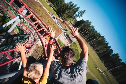Vater und Kind in einer Achterbahn mit erhobenen Händen, rote Schienen, Waldlandschaft im Hintergrund