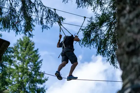 Person mit Helm und Klettergurt balanciert gesichert auf einem Drahtseil zwischen Baumwipfeln.