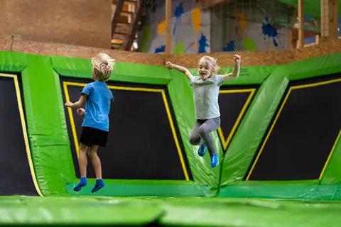 Zwei Kinder springen ausgelassen auf grünen Trampolinen in einer Indoor-Trampolinhalle.