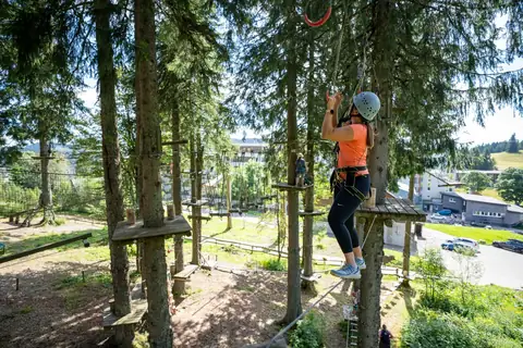 Frau mit Helm und Klettergurt balanciert auf einem Seil zwischen Baumplattformen im Hochseilgarten