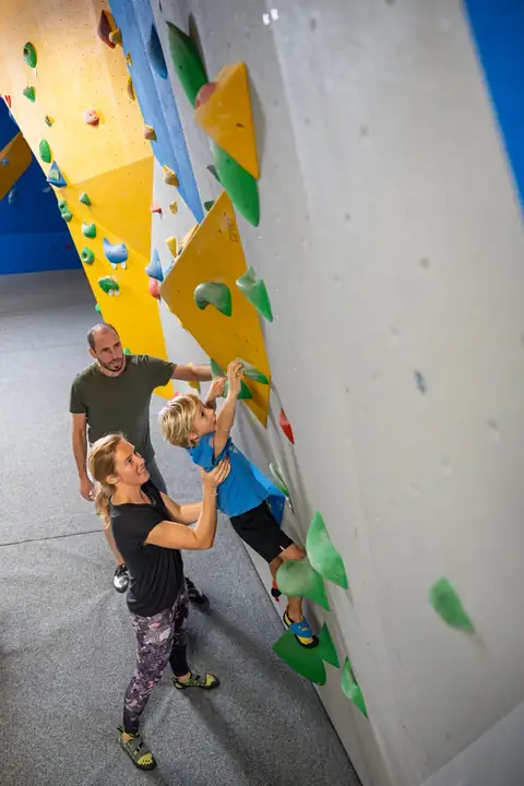 Kind klettert mit Unterstuetzung der Eltern an einer bunten Boulderwand in einer Kletterhalle