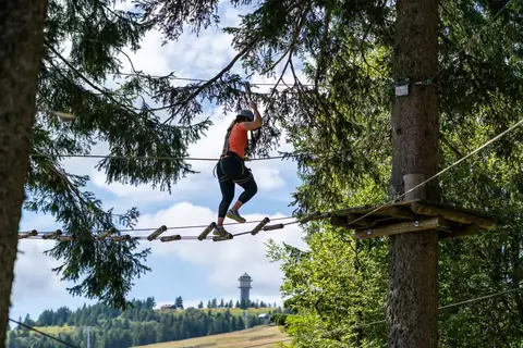 Frau überquert im Klettergurt einen Seilsteg im Hochseilgarten, im Hintergrund der Feldbergturm