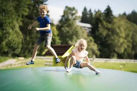 Zwei Kinder springen und rutschen lachend auf einem großen grünen Hüpfkissen im Freien