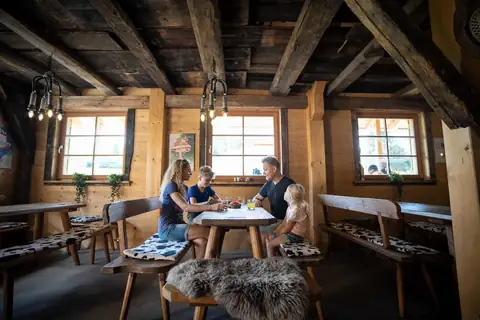 Familie sitzt bei Getränken an einem Holztisch in urgemütlicher Almhütte mit Kuhfell-Sitzkissen