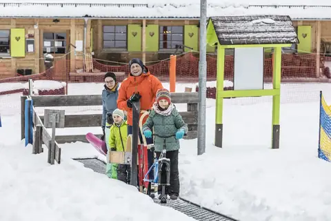 Familie mit Kindern zieht Schlitten und Laufrad über schneebedeckten Weg zur Rodelhütte