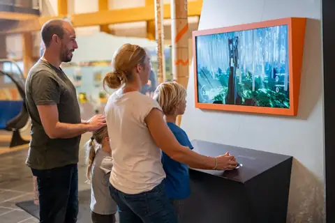 Familie an interaktiver Waldspiel-Station im Haus der Natur, Bildschirm zeigt Waldszene mit Harvester