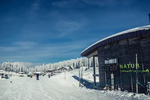 Verschneiter Vorplatz am Haus der Natur auf dem Feldberg, Holzfassade mit Schriftzug, dahinter Wintersportler und schneebedeckte Tannen