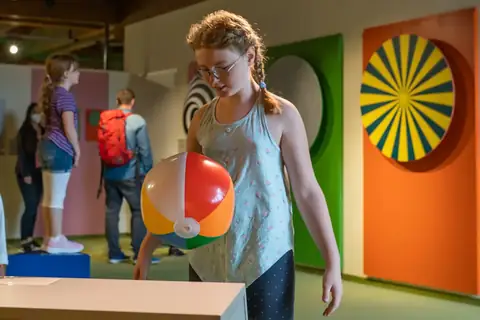 Mädchen mit Wasserball in bunter Mitmachausstellung mit Op-Art-Gemälden im Hintergrund