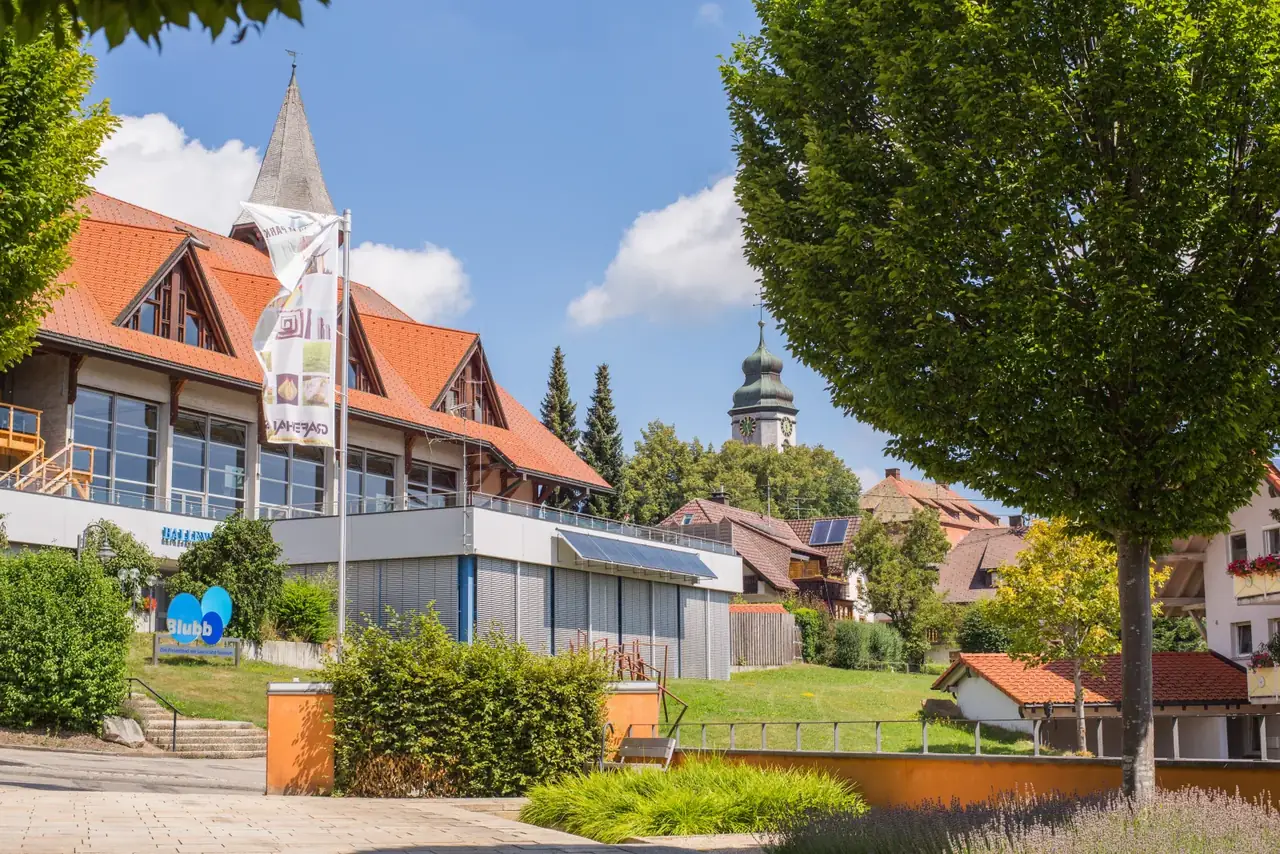 Hallenbad Blubb in Grafenhausen von außen, Wimpel am Eingang, dahinter Kirchturm und Ortsrand