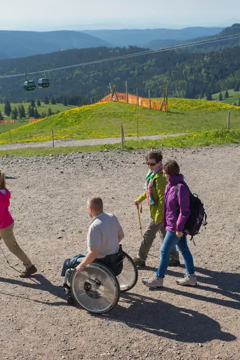 Rollstuhlfahrer und Wanderer auf Schotterweg am Feldberg mit Gondelanlage und Schwarzwaldpanorama