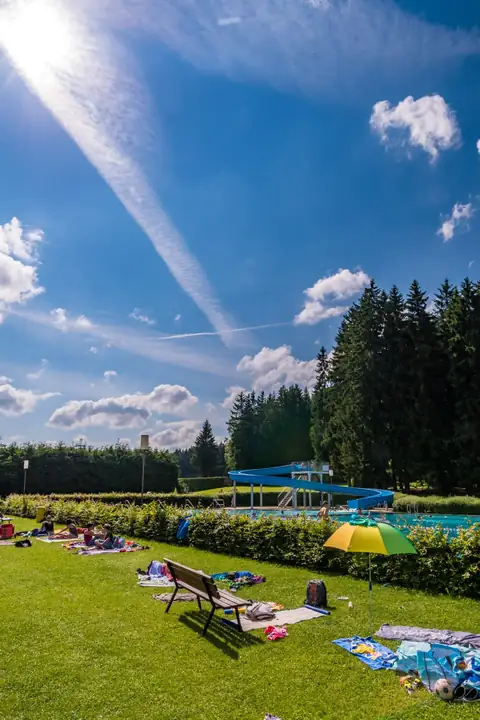 Freibad mit blauer Wasserrutsche und Schwimmbecken, im Vordergrund Liegewiese mit Sonnenschirm und Handtüchern