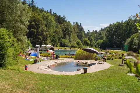 Naturbadesee mit kleinem Kinderbecken im Vordergrund, Liegewiese, Sonnenschirmen und bewaldeten Hängen im Hintergrund