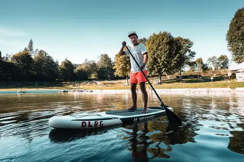Mann mit SUP-Board und Paddel steht auf einem Freibadsee, im Hintergrund Liegewiese und Bäume