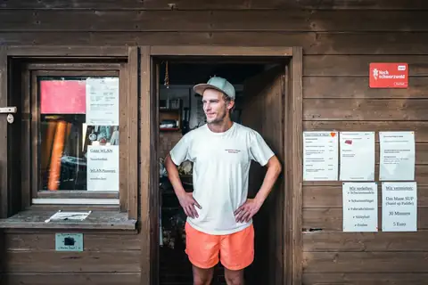 Mann in T-Shirt und oranger Badehose vor Holzhütte mit Info-Aushaengen, Freibad-Kiosk im Hintergrund